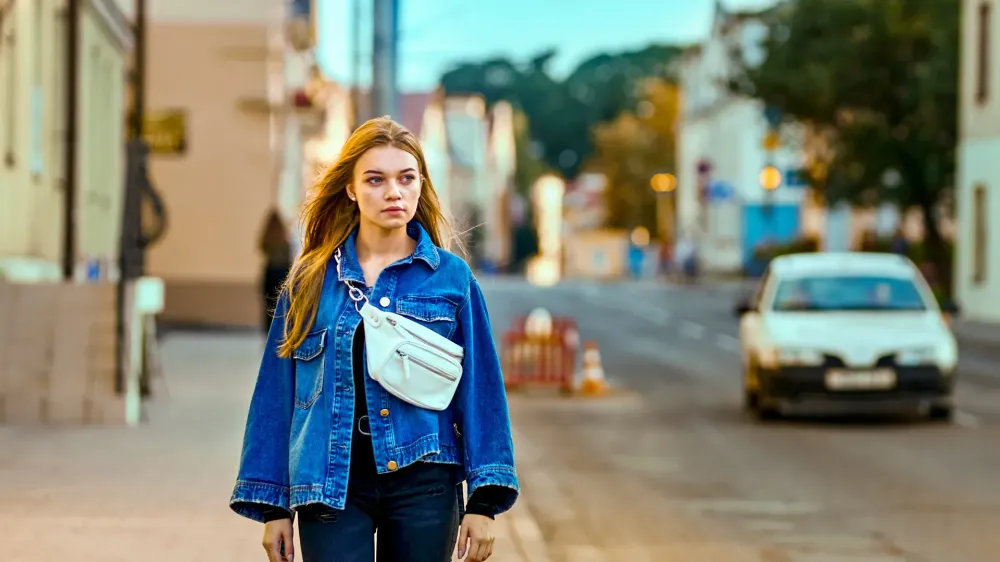 girl in a denim jacket and waist bag walks through the city along the road
